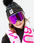 Person wearing a black knit hat with a pom-pom, pink goggles, and a pink 'Bunny' scarf on a white background
