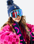 Person wearing a blue knit hat with a pom-pom, pink goggles, and a pink coat with a dark scarf on a white background