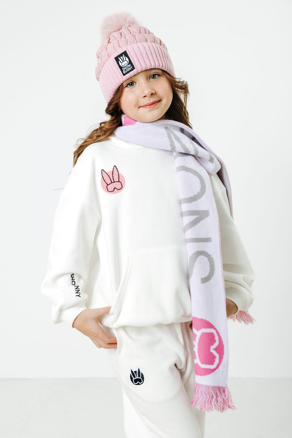 Child wearing a pink beanie and scarf with visible branding on a white background