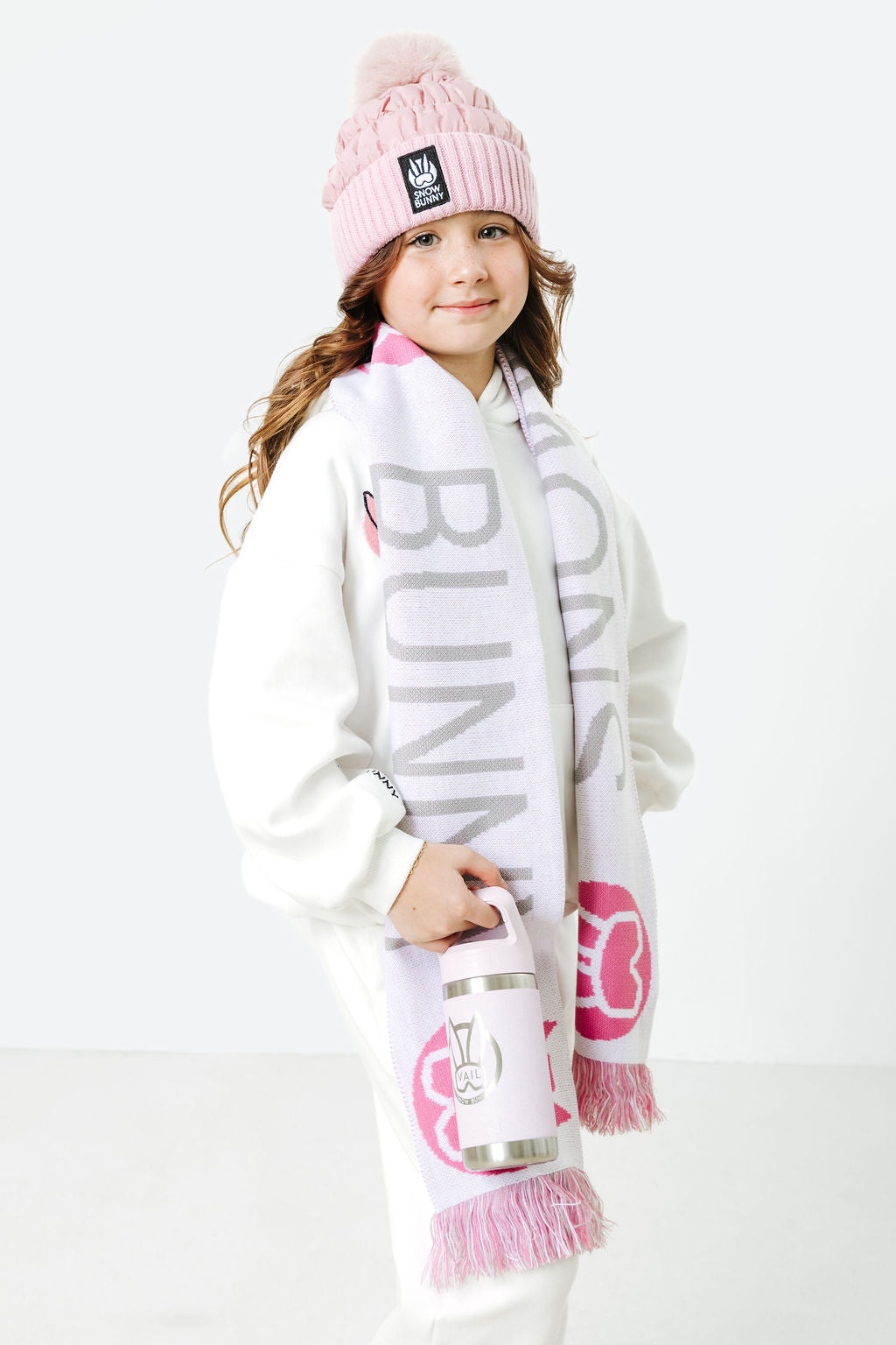Child wearing a pink beanie and scarf with visible branding on a white background