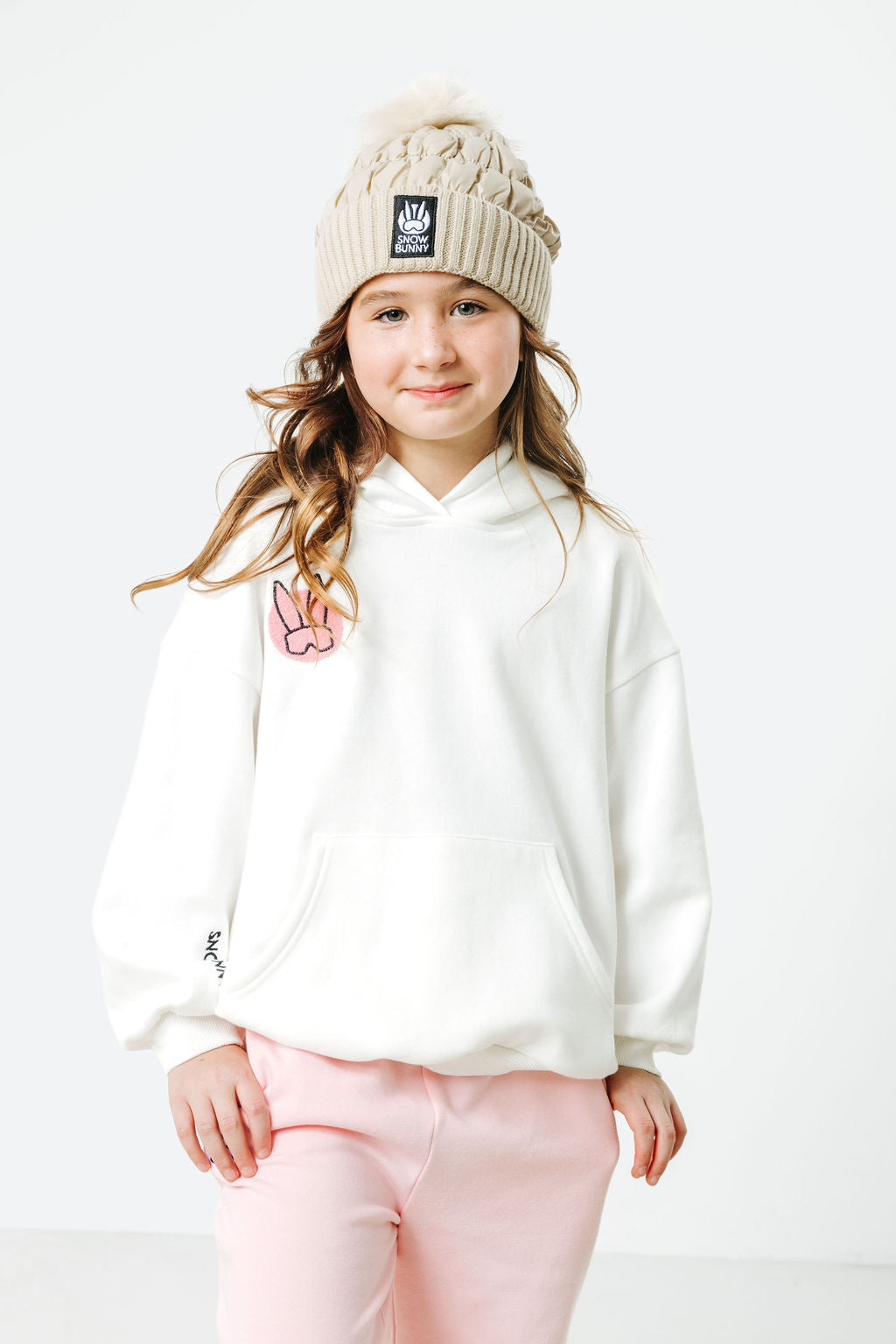 Young girl wearing a white hoodie and pink pants with a beige knit hat on a white background