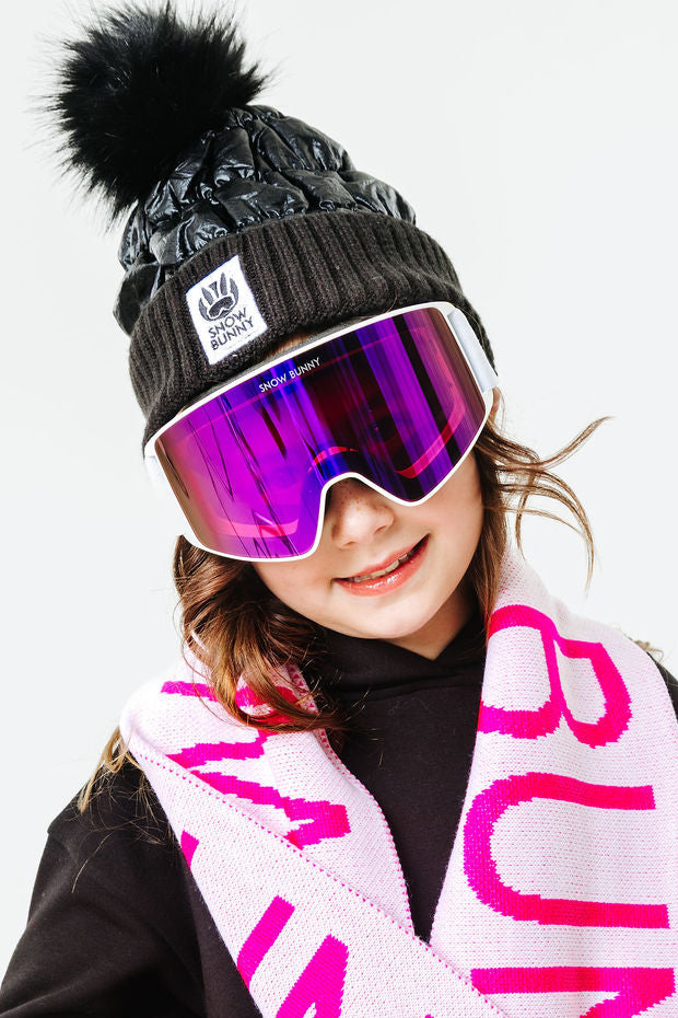 Person wearing a black knit hat with a pom-pom, pink goggles, and a pink 'Bunny' scarf on a white background