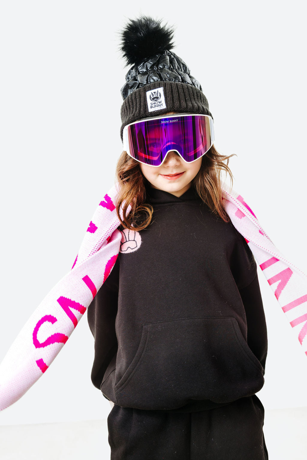 Person wearing a black beanie with a pom-pom, goggles, and a black sweatshirt with pink sleeves on a white background.