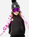 Person wearing a black beanie with a pom-pom, goggles, and a black sweatshirt with pink sleeves on a white background.