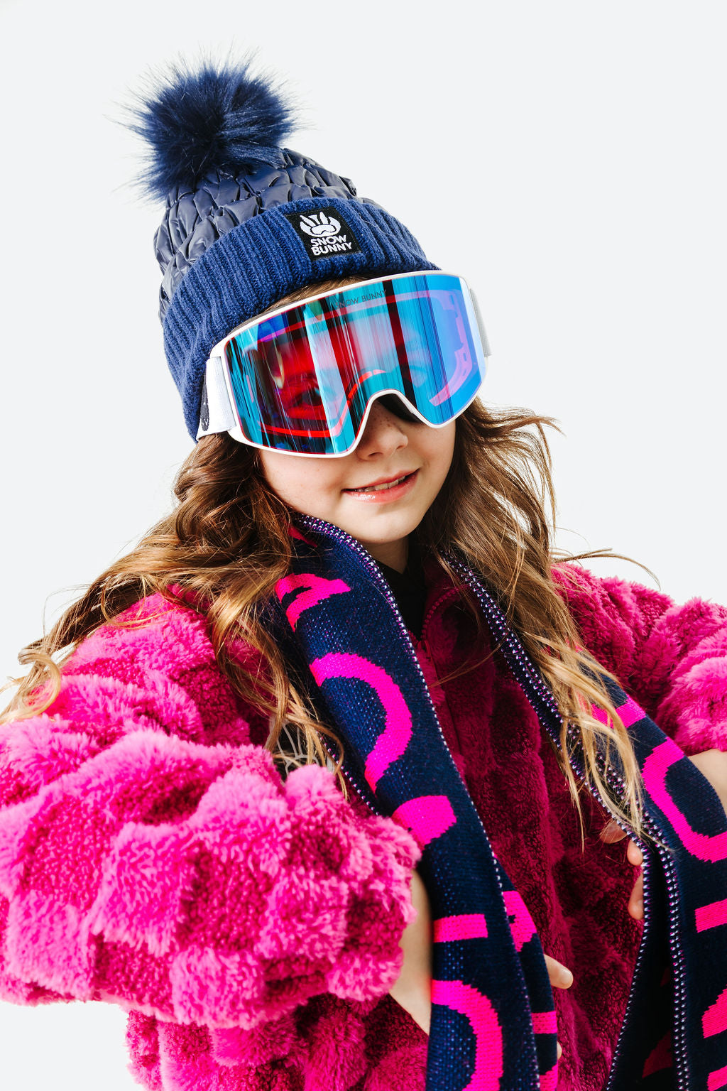 Person wearing a blue knit hat with a pom-pom, pink goggles, and a pink coat with a dark scarf on a white background
